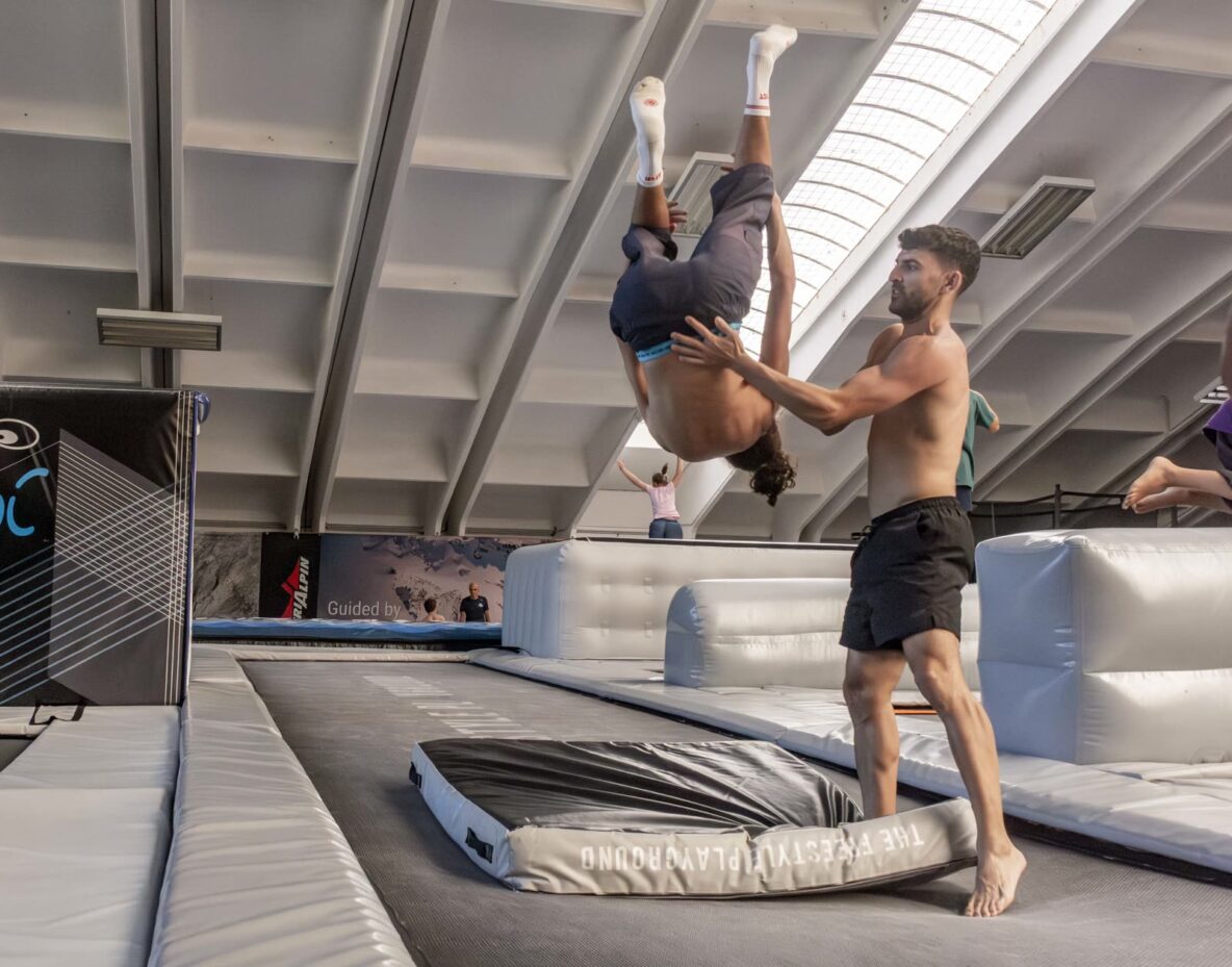 Un istruttore tiene le mani sulla schiena di un compagno a testa in giù, a metà di un salto all'indietro sul trampolino
