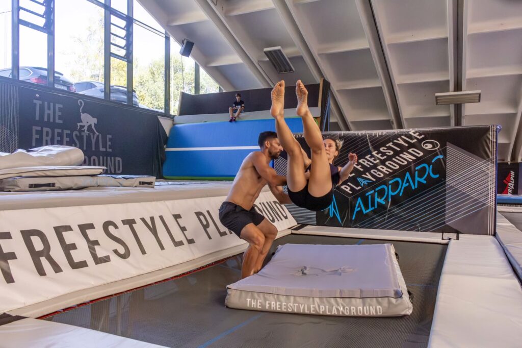 Un istruttore accompagna con le mani una compagna a metà di un salto all'indietro sul trampolino, sopra un materasso